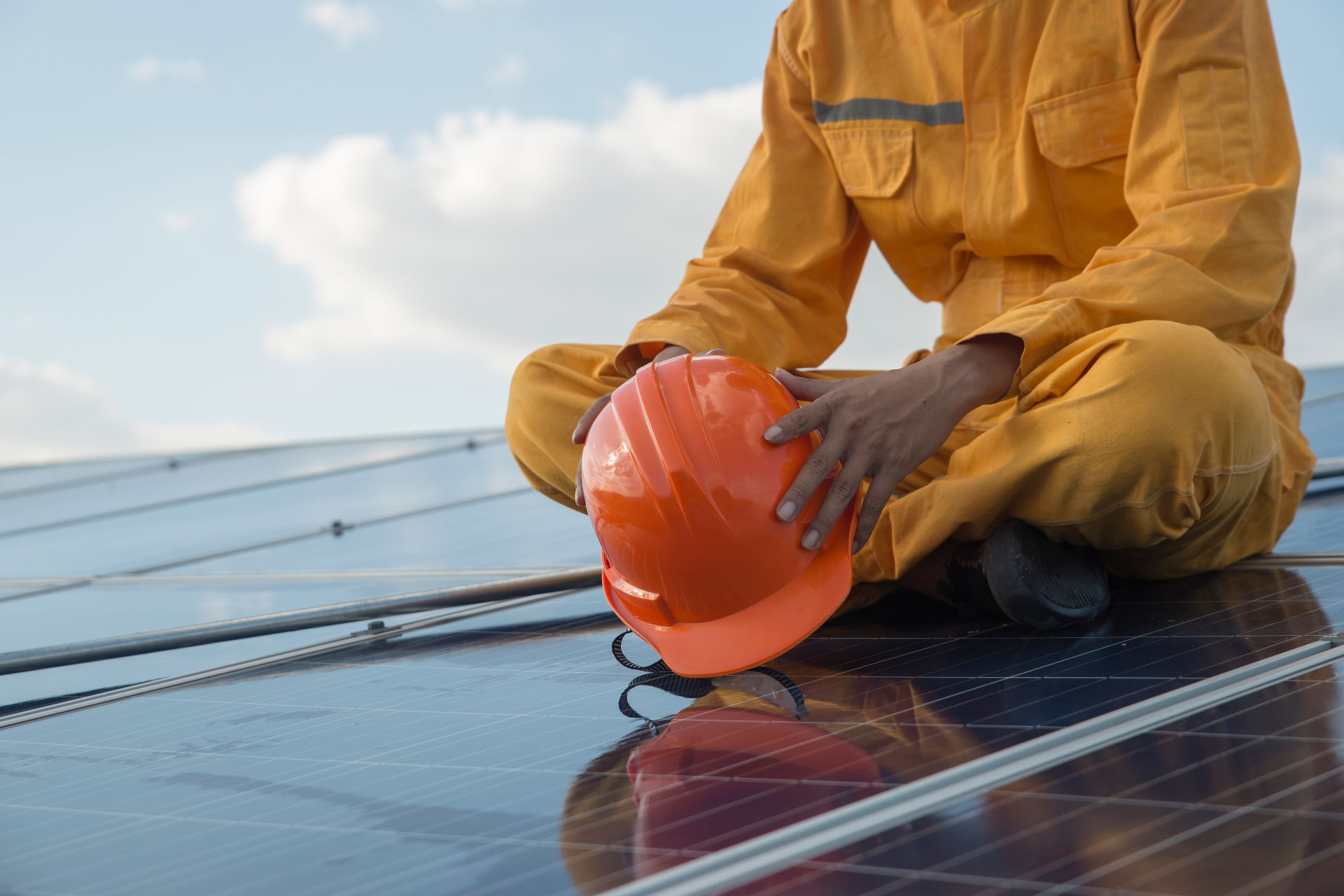 Technician in an orange jumpsuit sitting on a completed solar array holding a hardhat